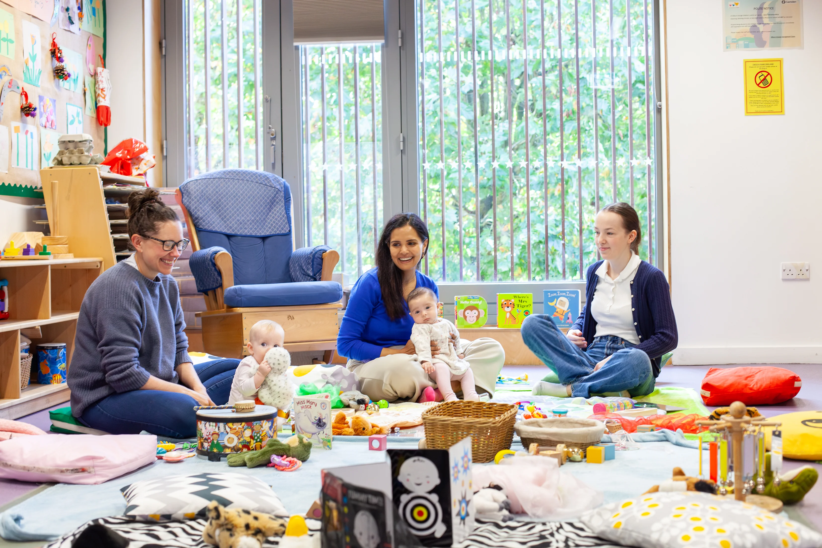 Three mothers and their babies sat on cushions with their babies playing with toys, pictured at a baby session.
