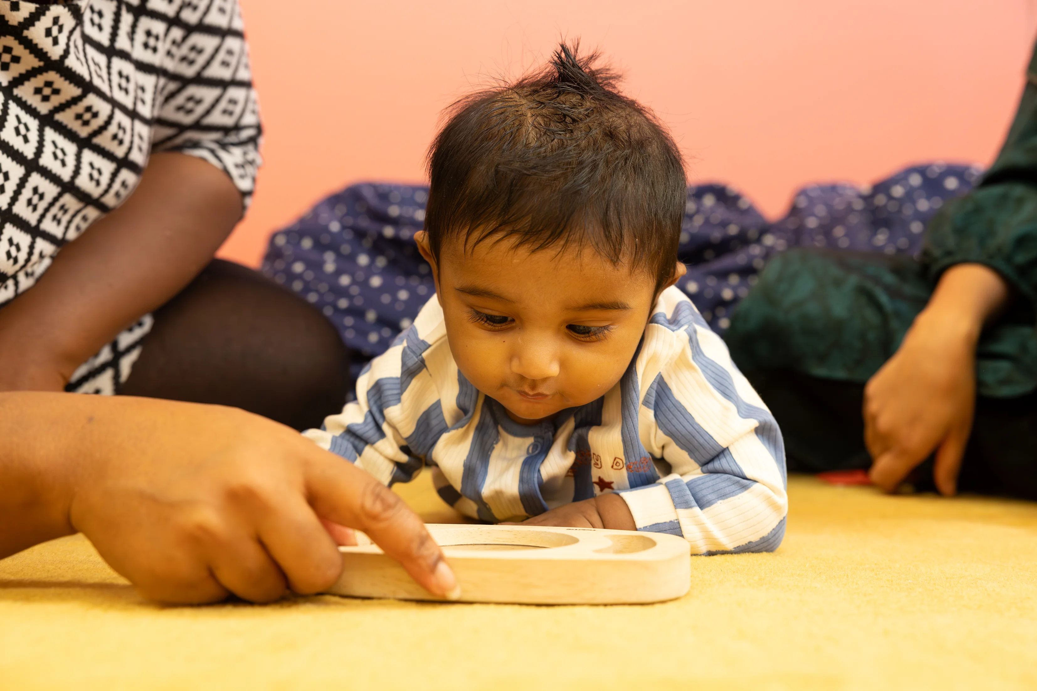 A baby lies on their tummy on a soft mat, focusing on a wooden toy as an adult points to it and encourages play.