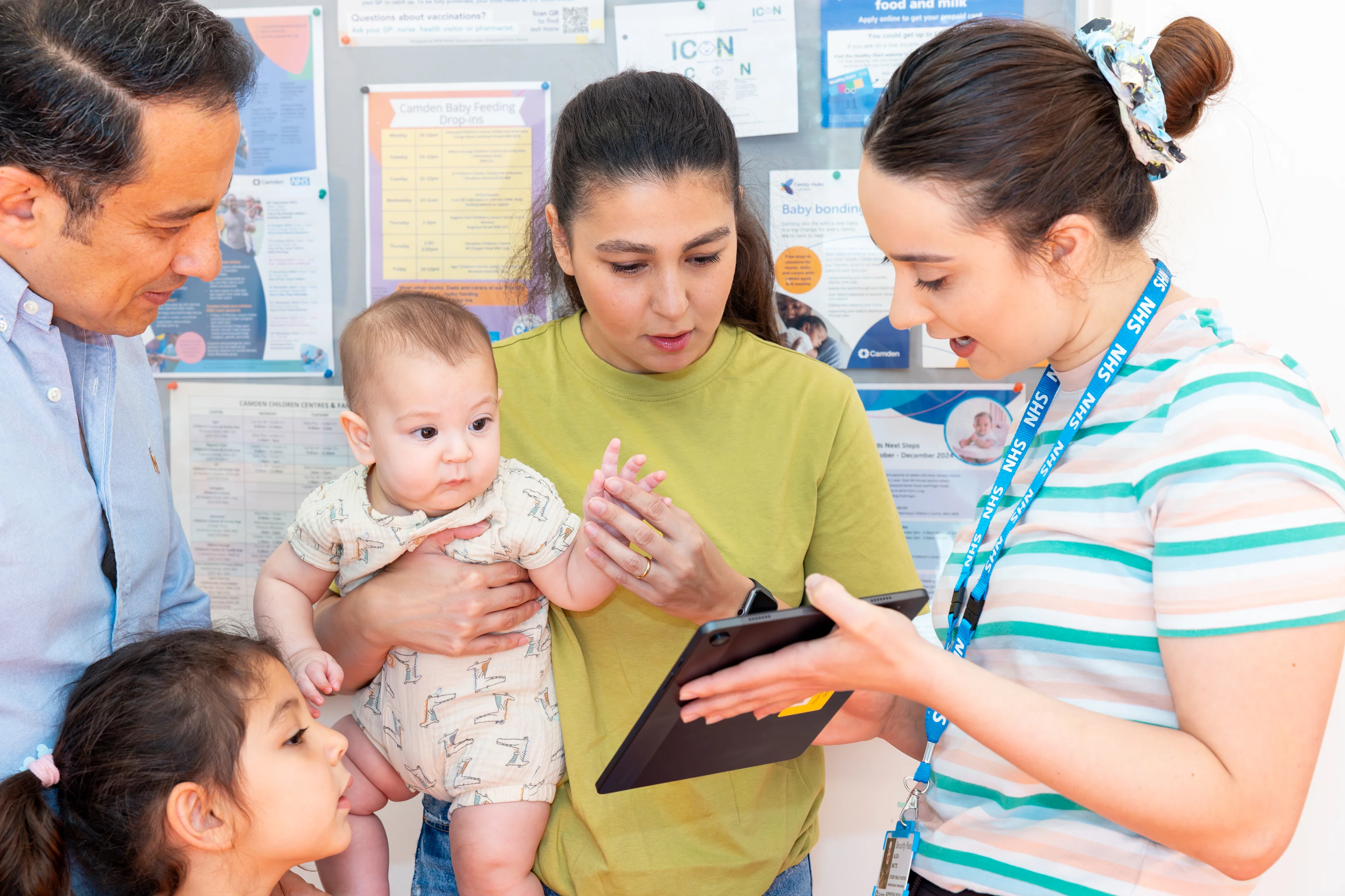 A healthcare worker shows information on a tablet to parents while one parent holds a baby and a young child looks on.