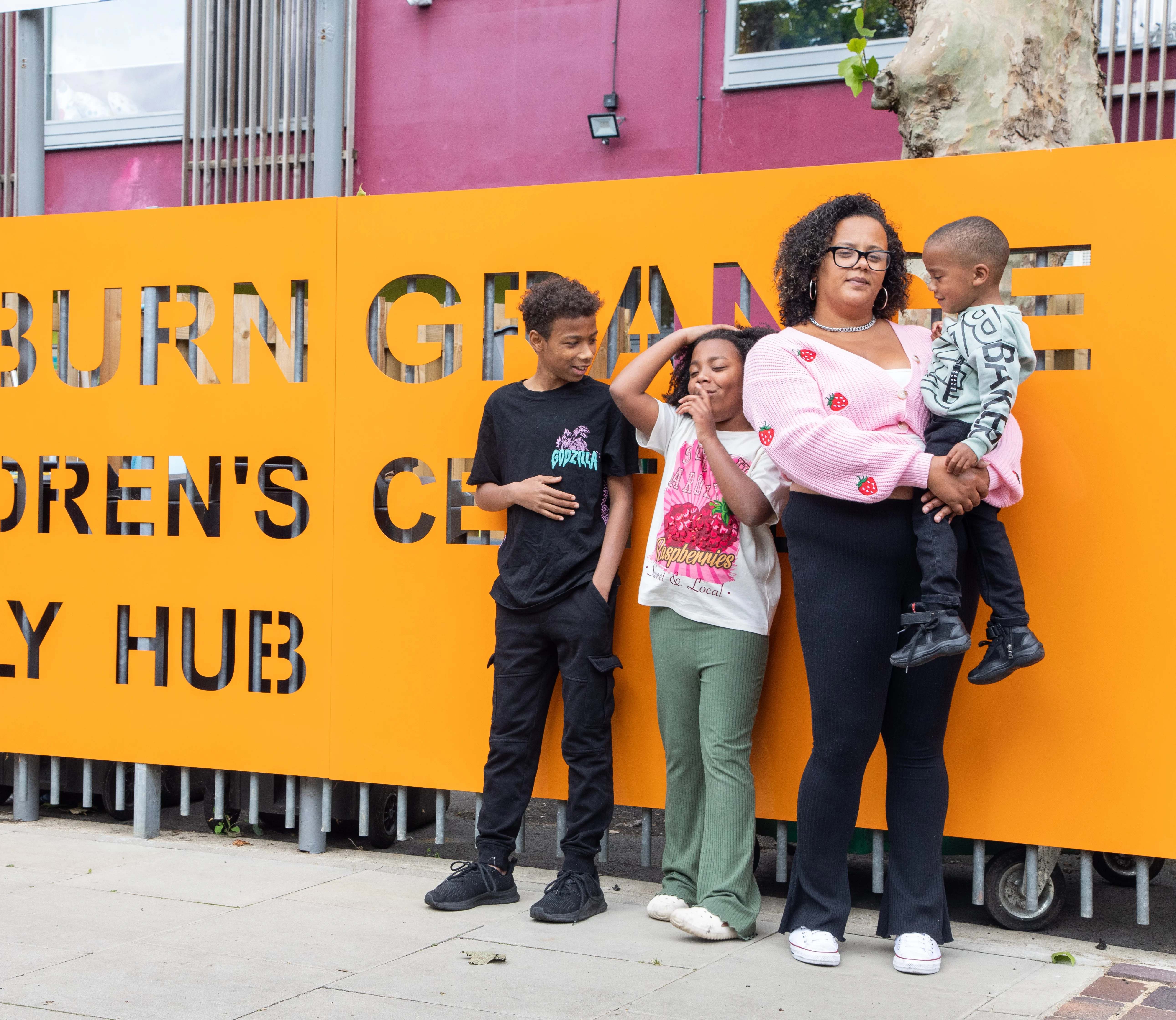 A mum and her 3 children of different ages standing outside Kilburn Grange Children's Centre and Family Hub.