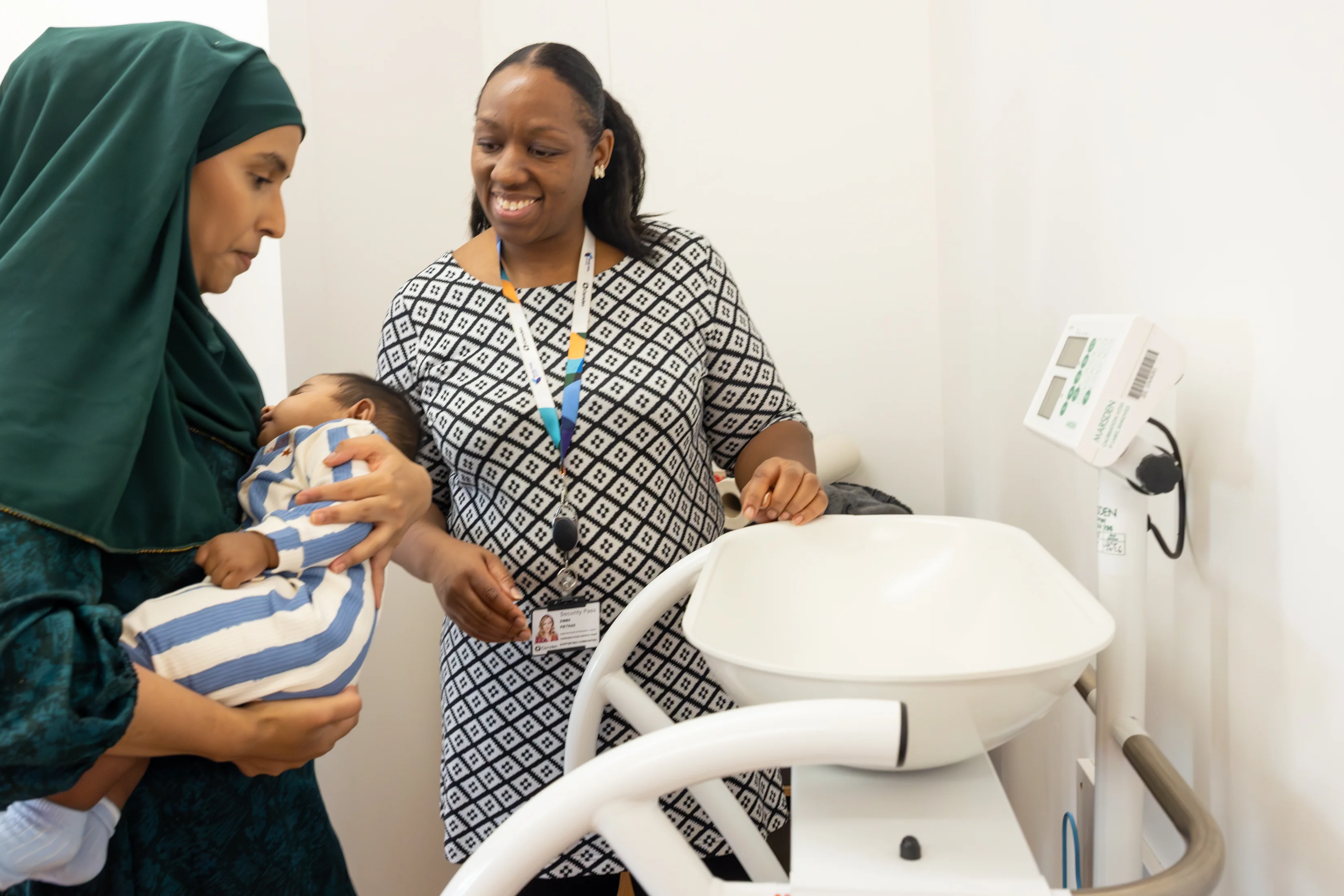 A healthcare worker stands beside a parent holding a baby, smiling as they support the baby being weighed on a scale in a clinic room.
